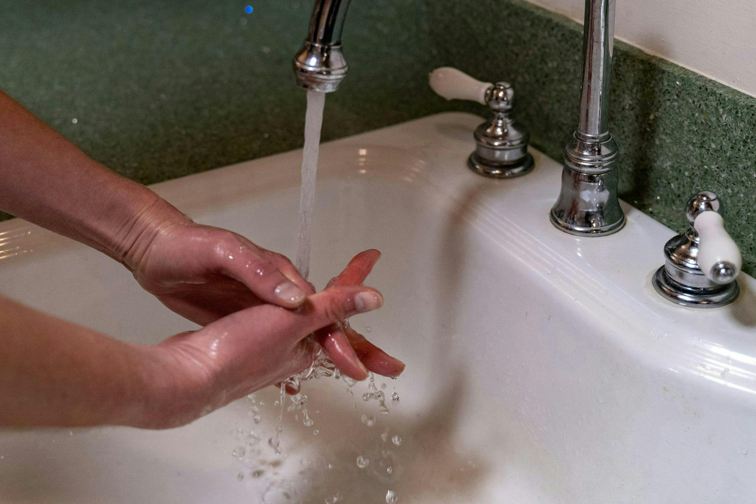 Hands scrub under a running faucet in a white sink, set within a green countertop. Water droplets splash as the tap flows.