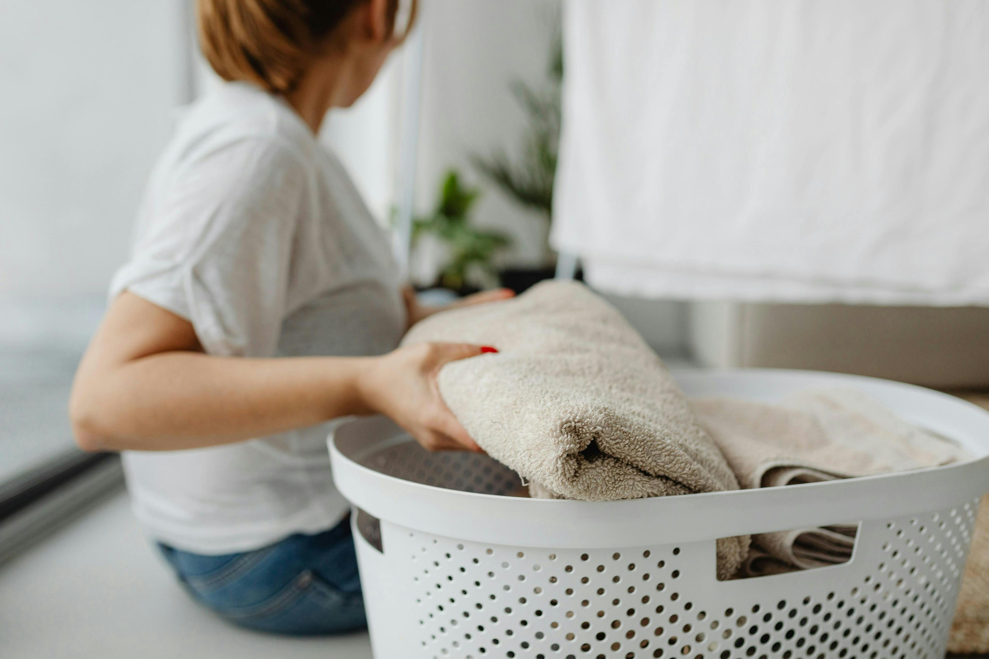 A person folding beige towels into a white laundry basket while seated indoors, with blurred plants and drying clothes in the background.