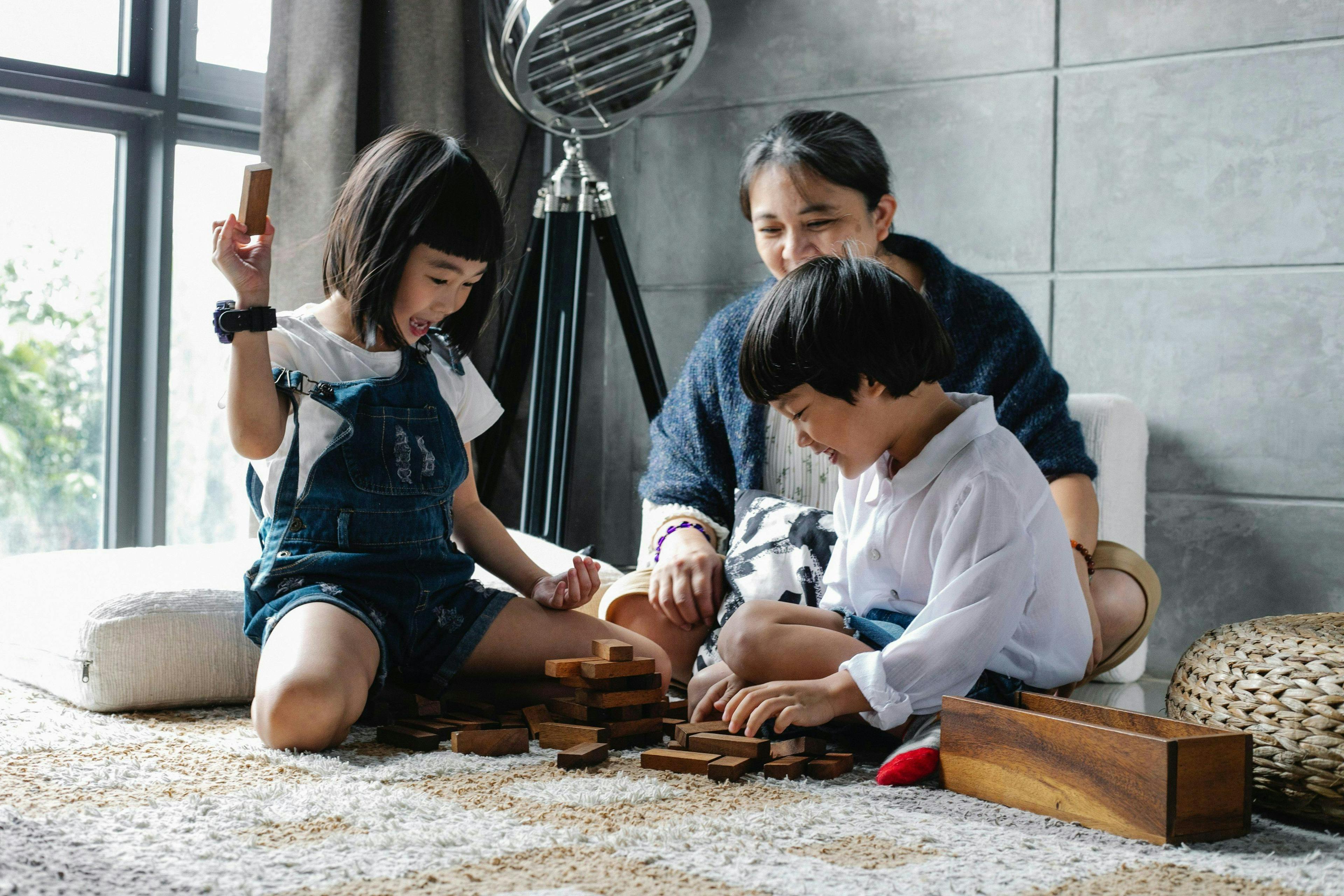 Two children are playing with wooden blocks while an adult woman observes and smiles; they are seated on the carpeted floor in a brightly lit, modern living room.