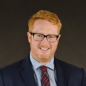 A man with red hair and glasses smiles while wearing a navy suit, striped shirt, and red plaid tie, set against a plain black background.