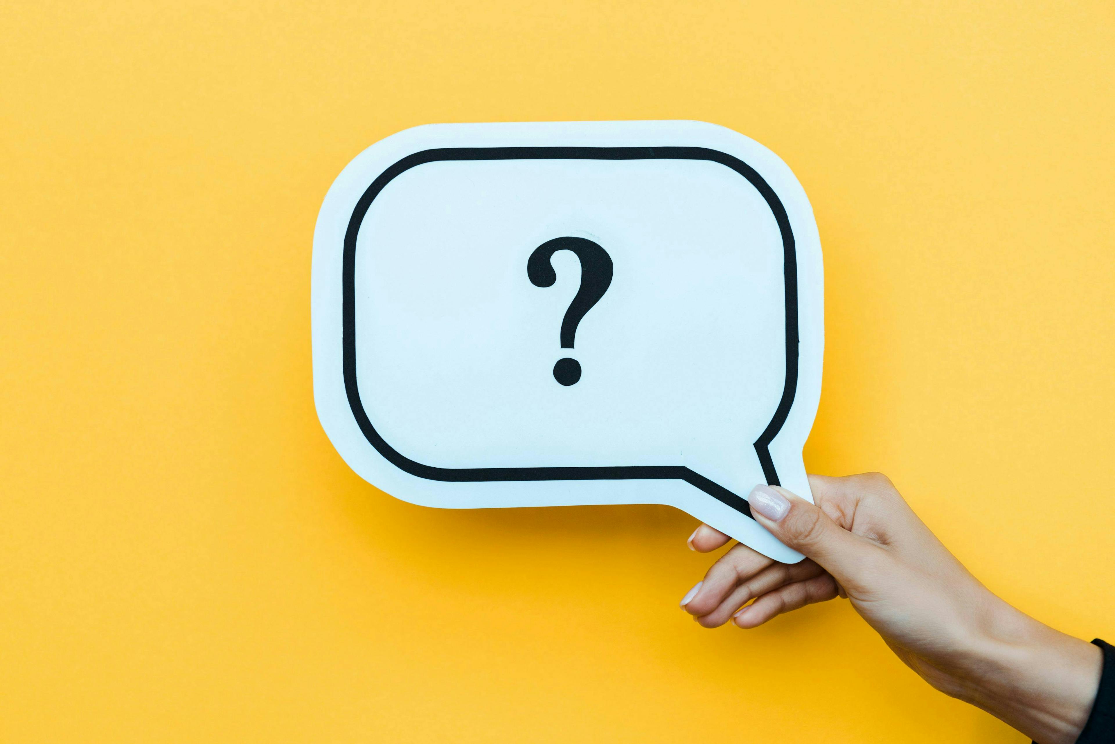 A hand holds a speech bubble cutout with a question mark, against a bright yellow background.