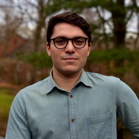 A person wearing glasses and a light blue buttoned shirt is standing outside with trees and blurred natural scenery in the background.