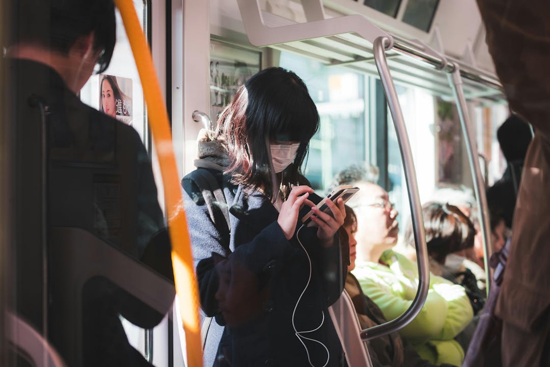 A person wearing a face mask uses a smartphone while standing in a crowded train with sunlight streaming through the windows. Other passengers are seated nearby.