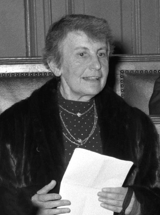 A woman holding a folded piece of paper is speaking in a formal setting, wearing a dark coat and patterned dress, with wood-paneled walls and a high-backed chair in the background.