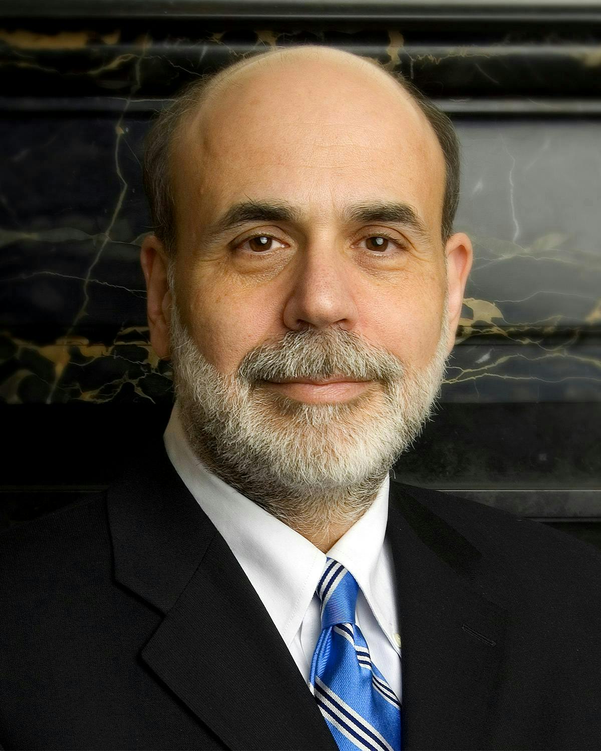 A bald man with a gray beard is wearing a suit and blue striped tie, looking directly at the camera against a dark marble background.