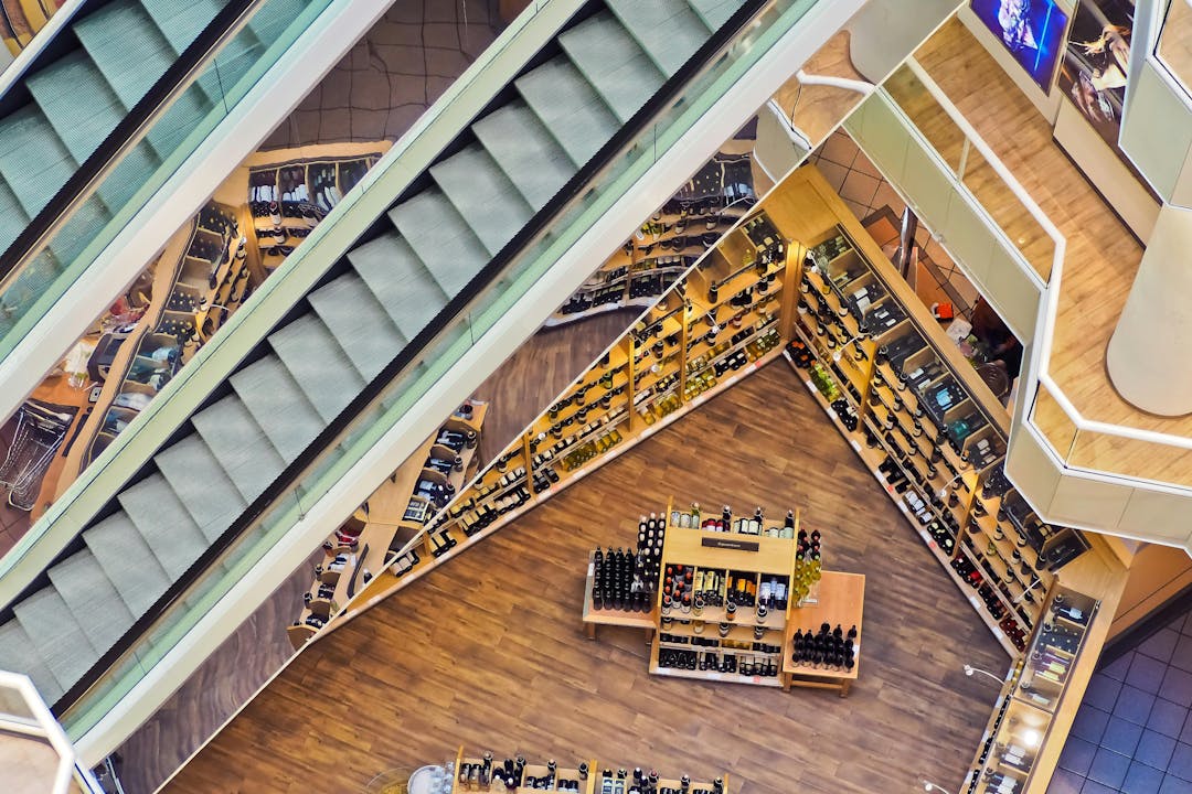Escalators descending above a wine shop with wooden flooring, surrounded by shelves of wine bottles in a multi-level shopping mall.
