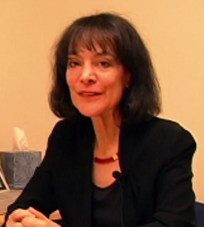 A woman with short dark hair sits at a table, wearing a black outfit. A tissue box is in the background on the left against a plain wall.