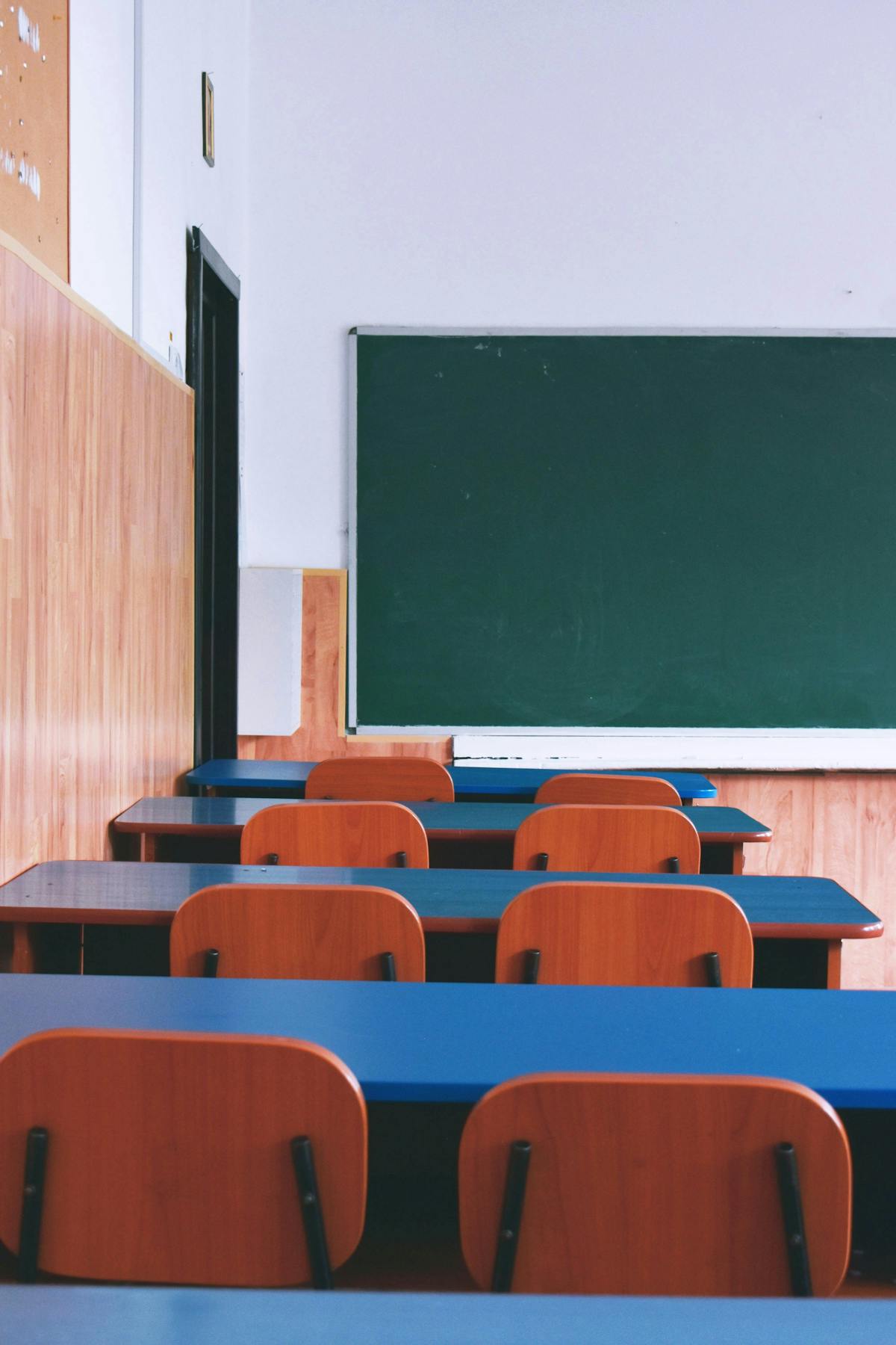 Rows of empty wooden desks face a green chalkboard in a well-lit classroom with wood-paneled walls.