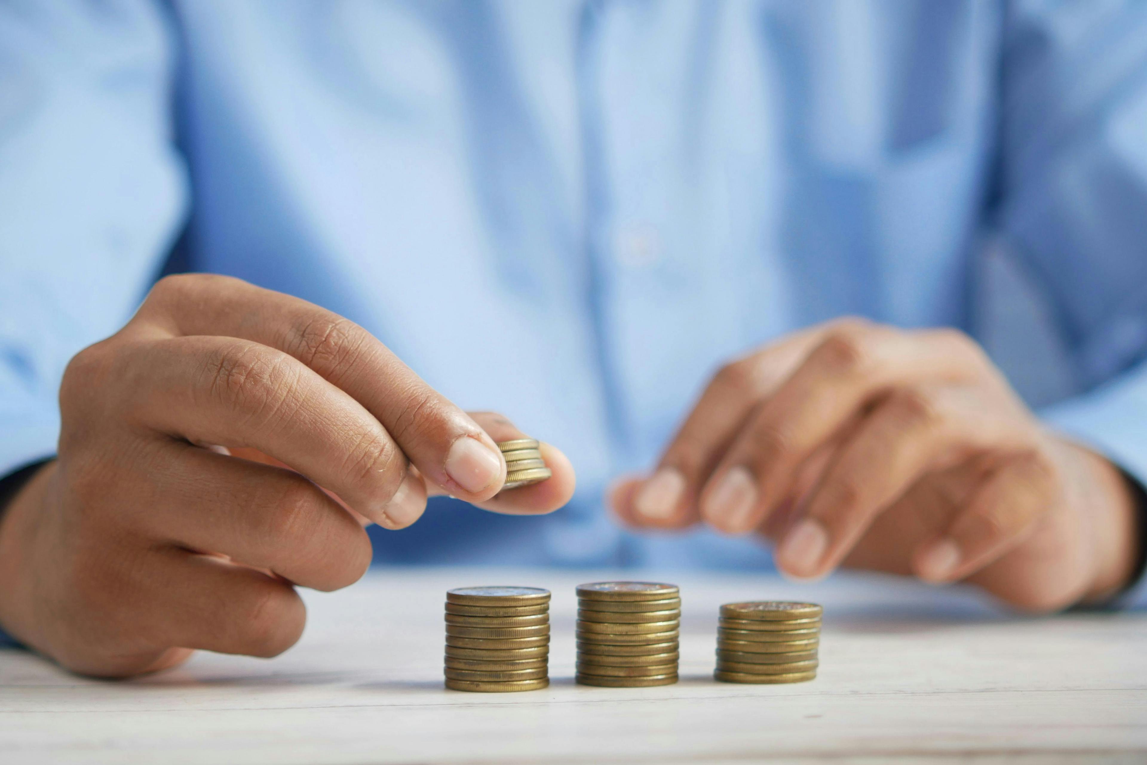 Hands stack coins on a white table, arranging three columns; the person wears a blue shirt, suggesting a context of financial planning or saving.