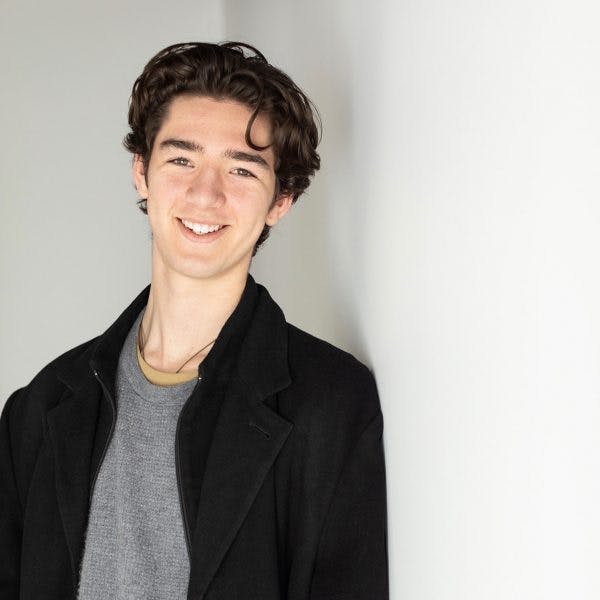 A young man, wearing a dark coat and gray shirt, smiles while leaning against a white wall in a brightly lit room.