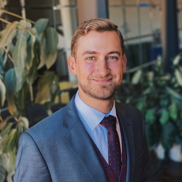 A man in a blue suit and red tie smiles while standing indoors, surrounded by office plants.