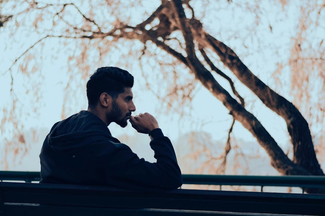 A man sits on a bench, resting his chin on his hand, thoughtfully gazing forward. The backdrop features bare-branched trees, suggesting a fall or winter setting.