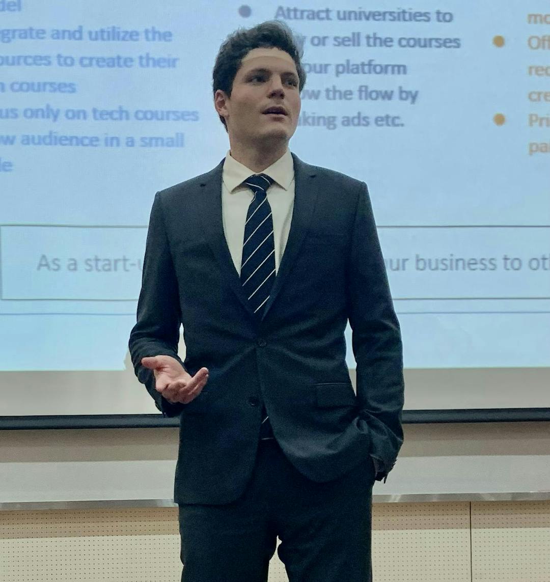 A man in a suit and tie stands and gestures while presenting in front of a projected screen displaying text and bullet points inside a lecture hall or conference room.