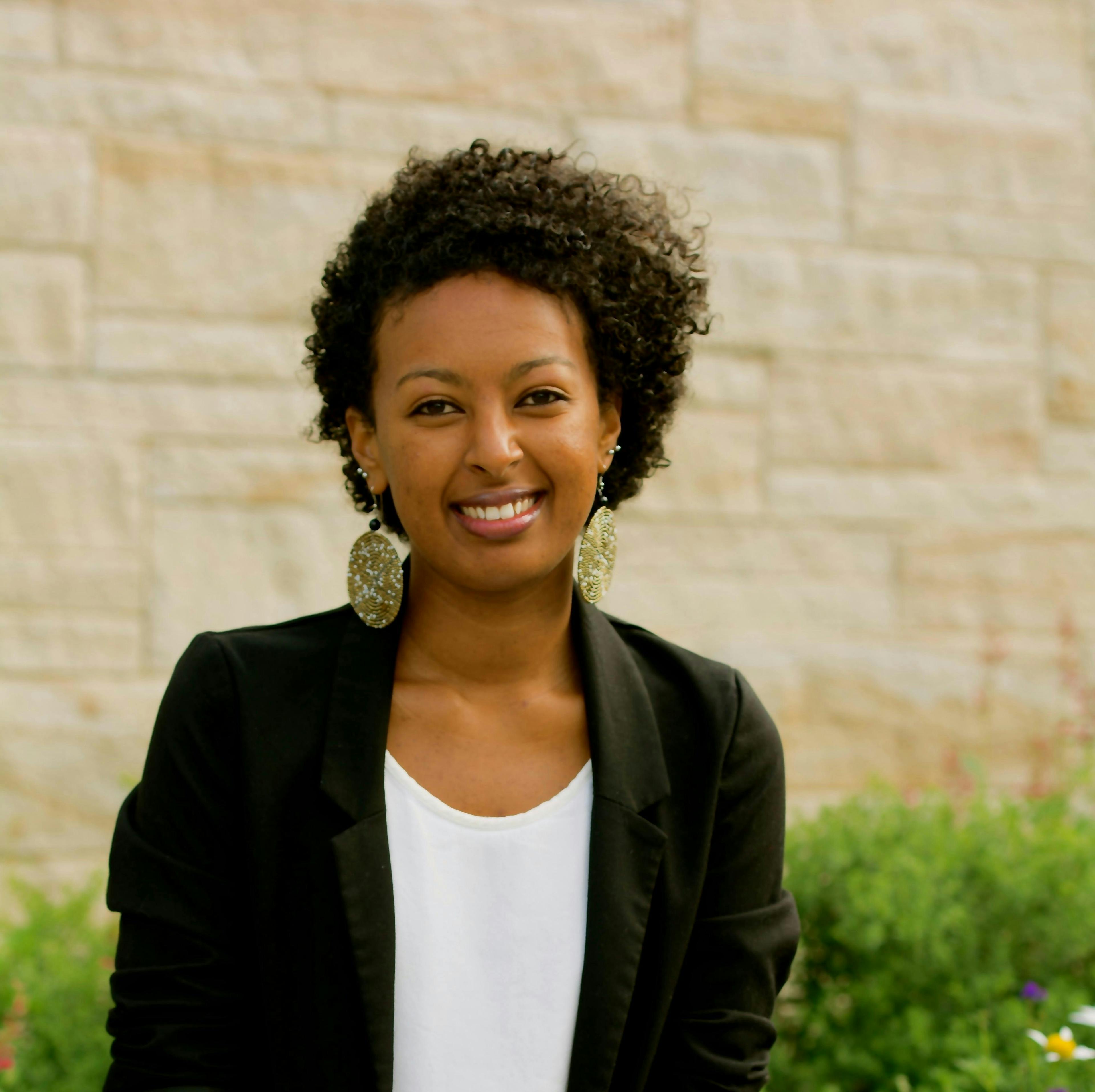 A person with curly hair, wearing a dark blazer and large earrings, smiles while standing in front of a beige stone wall and greenery.