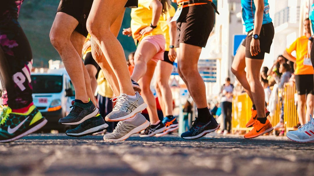 Runners in colorful athletic shoes stride on a sunlit street during a race, surrounded by spectators and parked vehicles, indicating a lively outdoor event.
