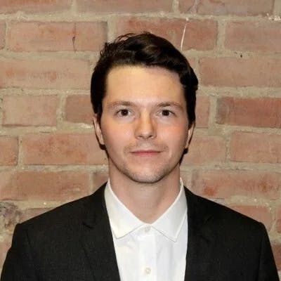 A person stands against a brick wall, wearing a dark suit jacket over a white button-up shirt, looking directly ahead.