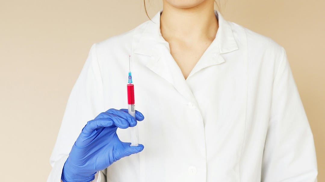A person in a white lab coat holds a syringe with red liquid using a gloved hand, set against a plain background.