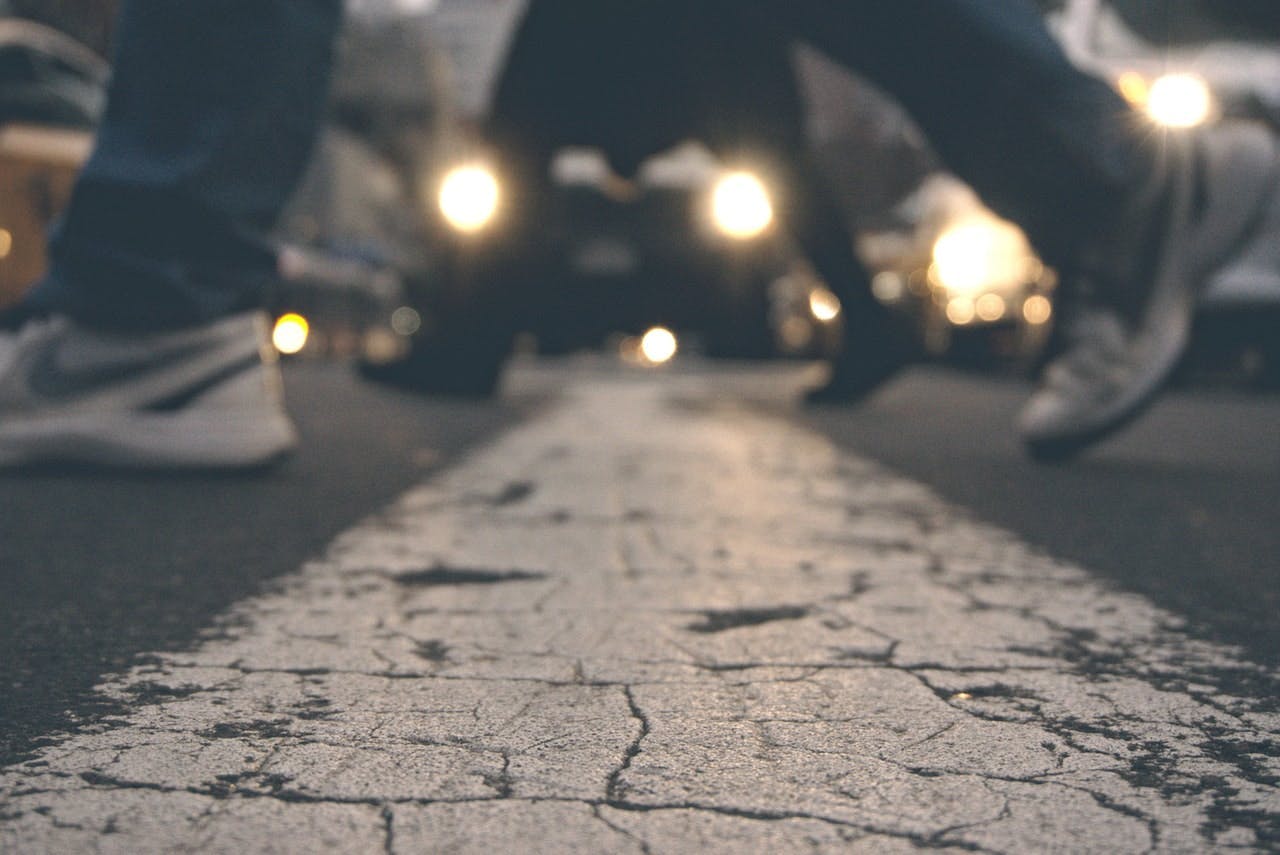 Shoes walk across a cracked white line on a dimly-lit street, with blurred headlights from approaching vehicles in the background.