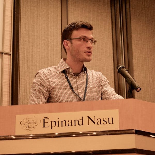 A man speaks at a podium labeled "Épinard Nasu" with a microphone, in a conference room with beige panel walls. The sign also includes "Hotel Épinard Nasu".