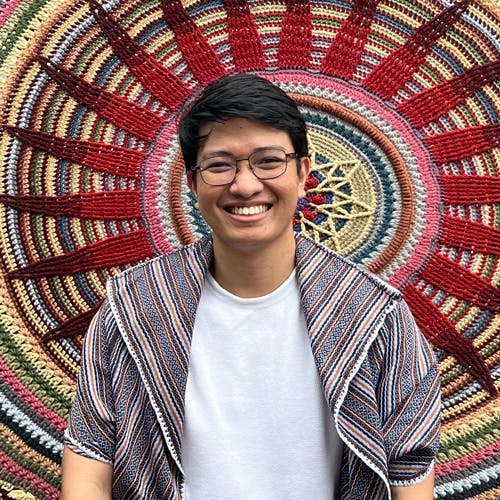 A person smiles while standing in front of a colorful, circular woven tapestry with intricate patterns, predominantly featuring red, green, and beige colors. The person wears glasses, a white shirt, and a striped outer garment.