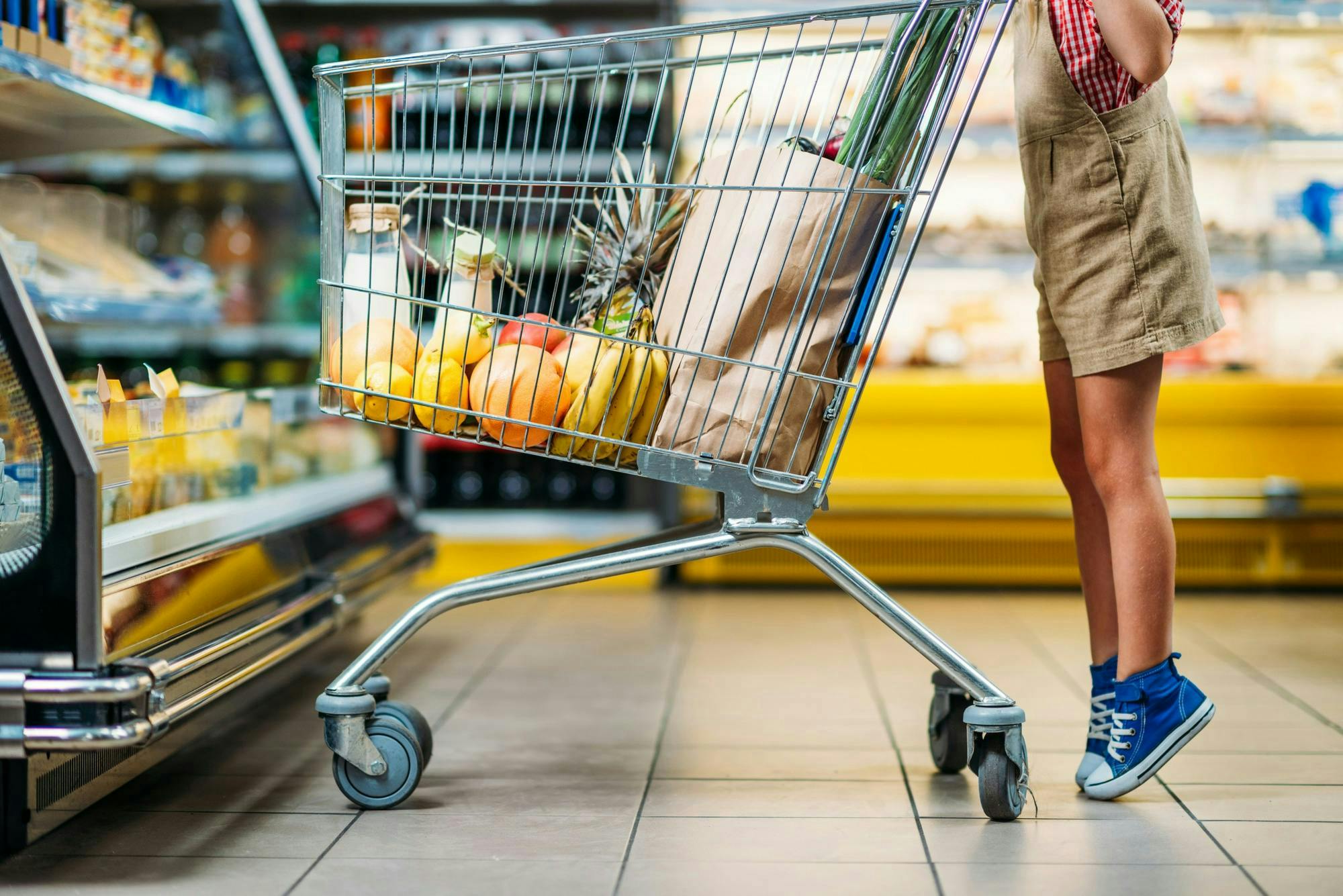 A shopping cart filled with groceries, including fruits and a paper bag, is pushed by a child wearing blue sneakers and overalls in a grocery store aisle.