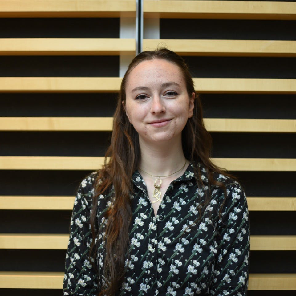 A person is smiling, standing in front of a wall with horizontal wooden slats. They wear a dark, patterned shirt and a necklace with a pendant.