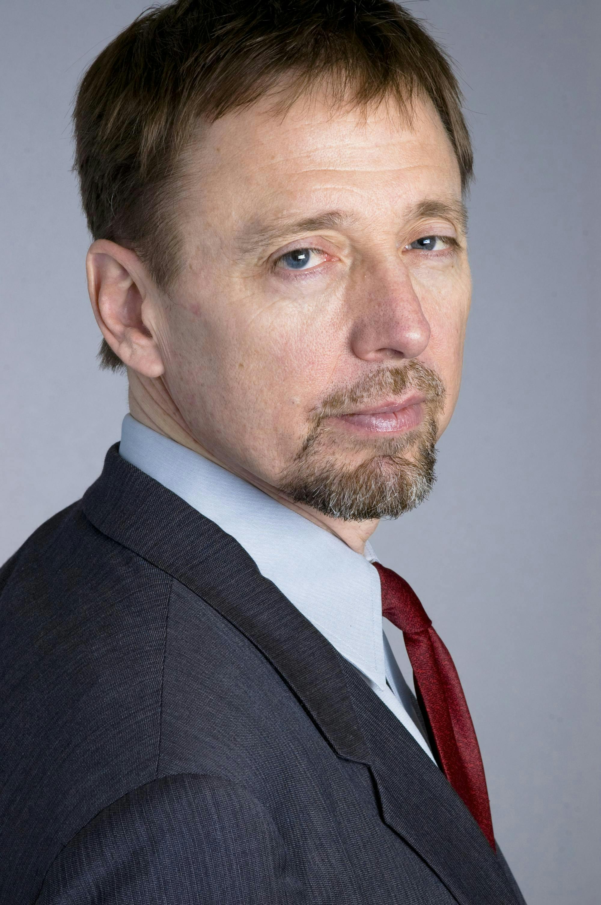 Middle-aged man with short brown hair and a goatee wearing a suit, white shirt, and red tie, facing the camera with a serious expression against a plain gray background.