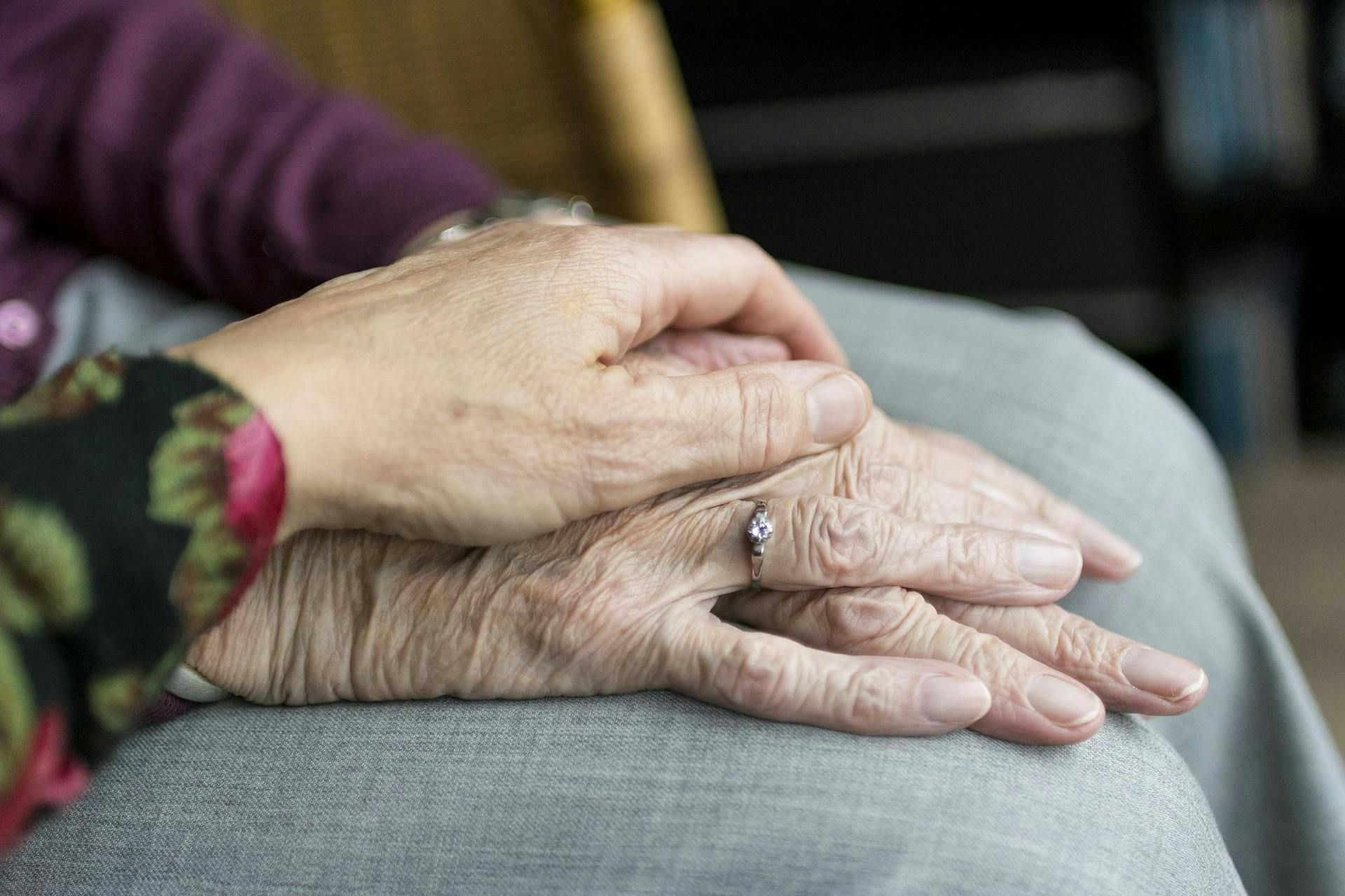 An elderly person's hand with a ring is clasped gently by another person's hand, set against a background of gray fabric and blurred room details.