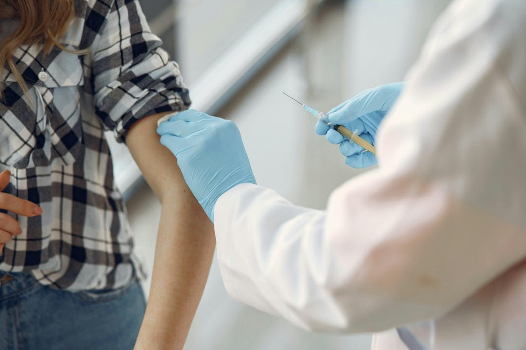 A person in a plaid shirt receives an injection from a healthcare professional wearing blue gloves in a clinical setting.