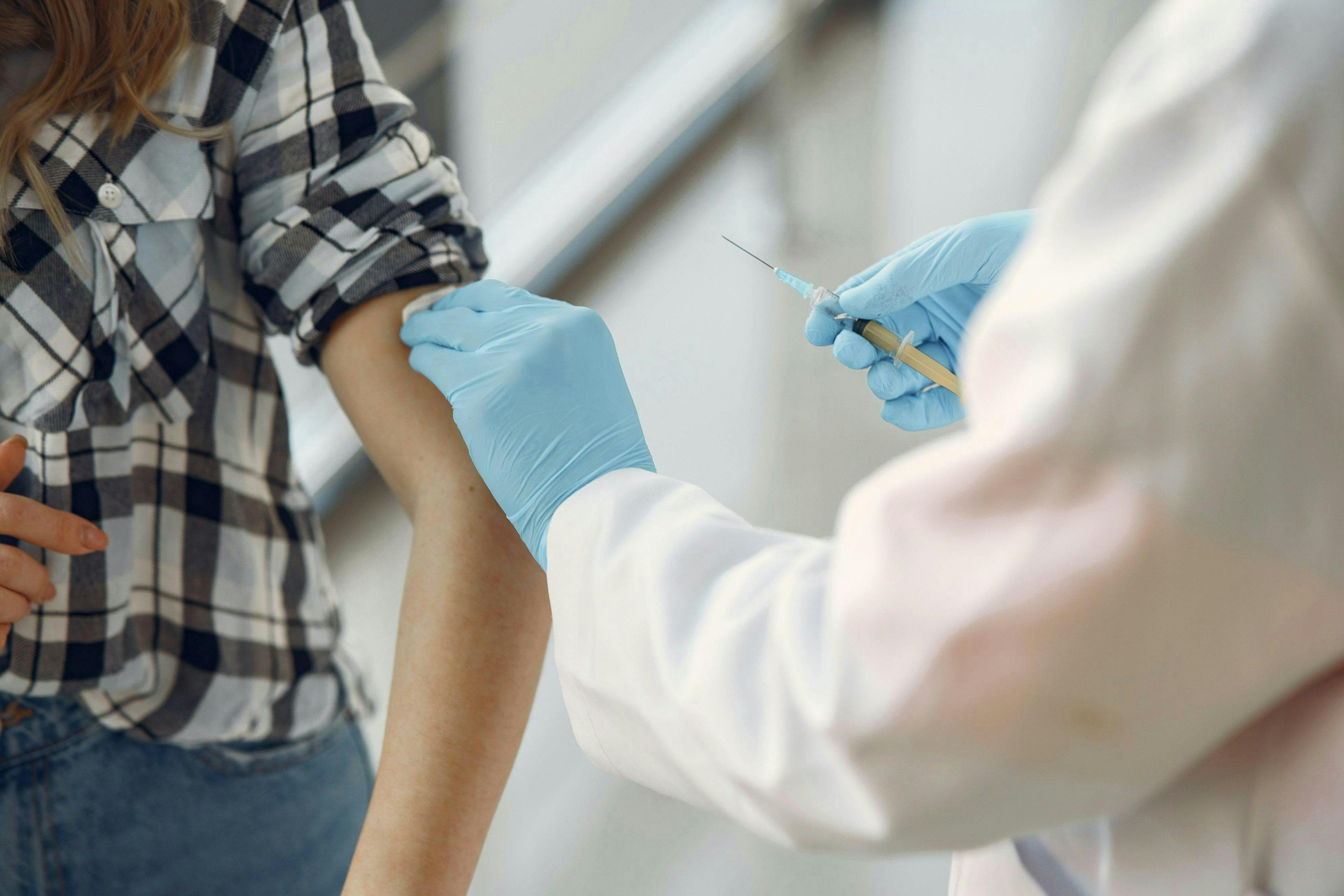 A person in a plaid shirt receives an injection from a healthcare professional wearing blue gloves in a clinical setting.
