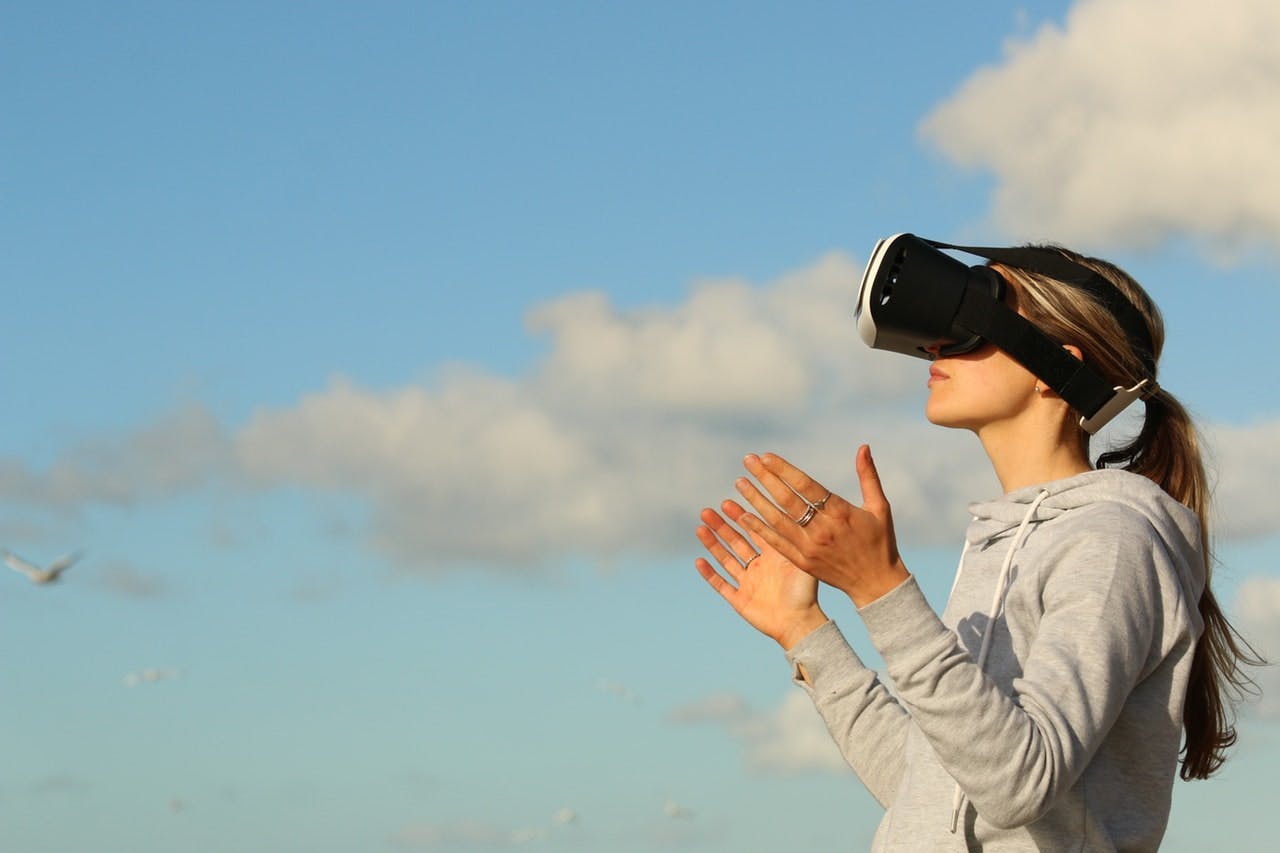 Person wearing a virtual reality headset, gesturing with hands, standing outdoors under a clear blue sky with scattered clouds and a few seagulls flying in the background.