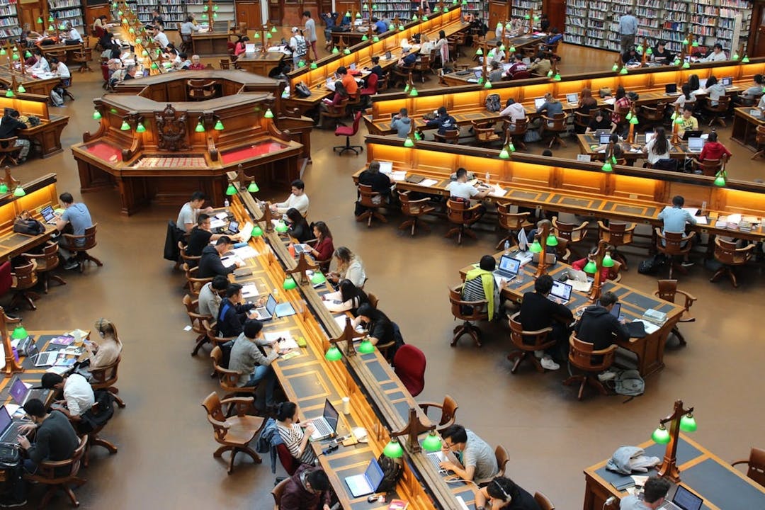 Students are studying at long, wooden tables in a large, well-lit library with green desk lamps and surrounded by bookshelves. The space is busy and quiet.