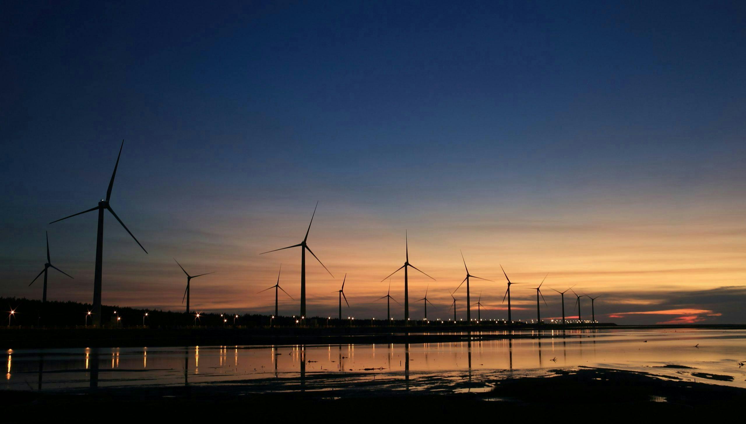 Wind turbines standing still, silhouetted against a dramatic sunset sky, are lined up along a coastal shoreline reflecting on calm water.