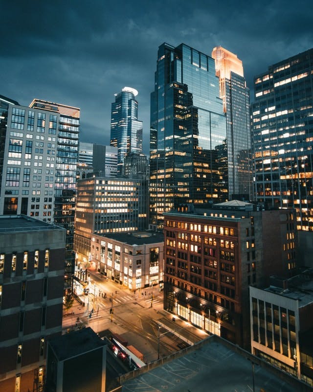 Skyscrapers with lit windows stand tall under a dark, cloudy sky; streetlights illuminate the empty streets below in a bustling urban cityscape.