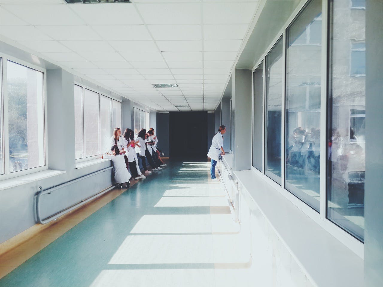 Eight people in white coats lean against a sunlit hallway's large windows, while one person stands on the right side.
