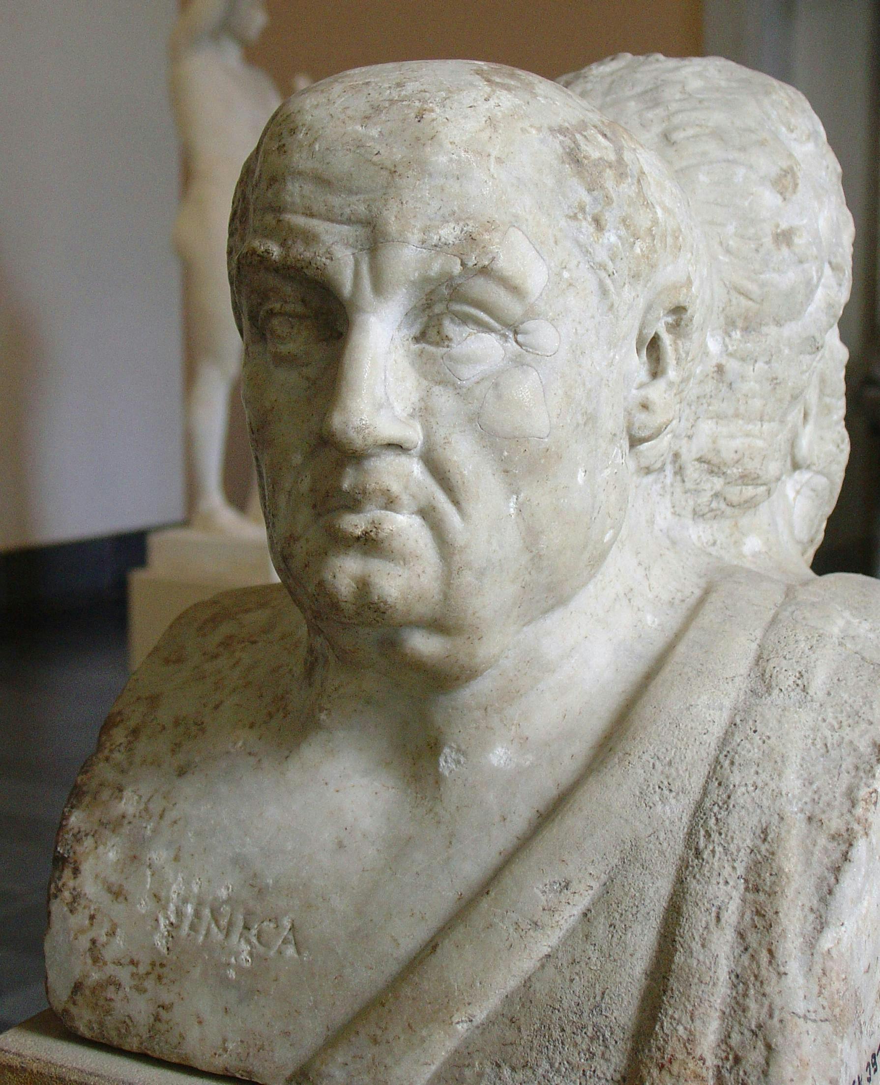 Marble bust portraying an older man with a serious expression in a museum setting, showing partially visible inscription "NIRKA" on the chest; blurred statues are in the background.