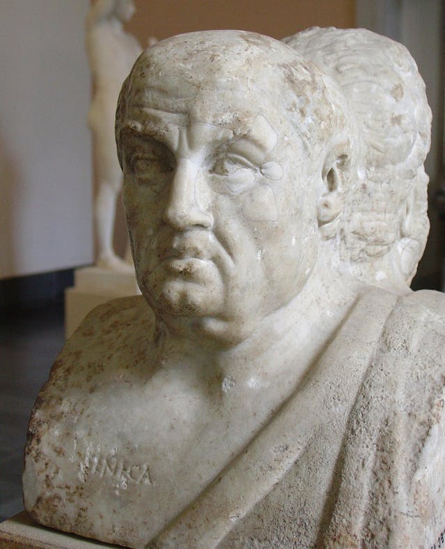 Marble bust portraying an older man with a serious expression in a museum setting, showing partially visible inscription "NIRKA" on the chest; blurred statues are in the background.
