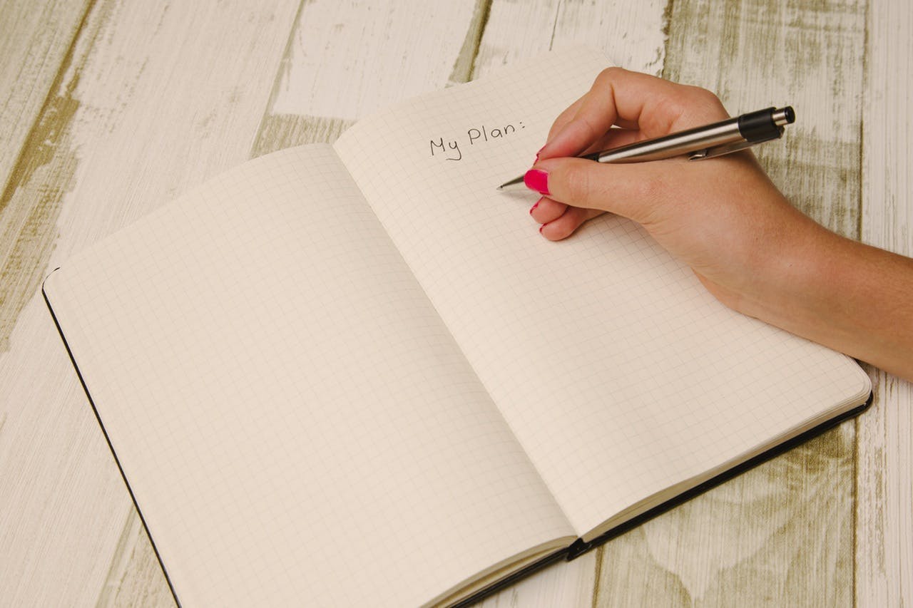 A hand with pink nail polish writes "My Plan:" in a grid notebook using a silver pen, on a wooden table with a rustic finish.