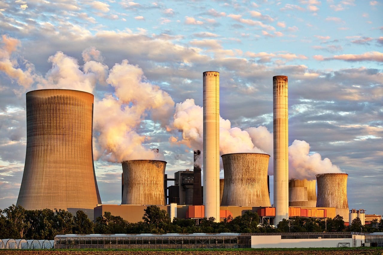 Several cooling towers and smokestacks emit steam and smoke in an industrial area, set against a partly cloudy sky with greenery in the foreground.