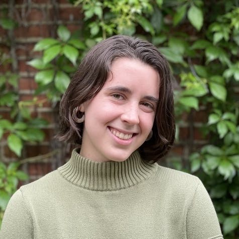 A person with short brown hair smiles, wearing a green turtleneck sweater. Behind them is a brick wall partially covered with green leaves and vines.