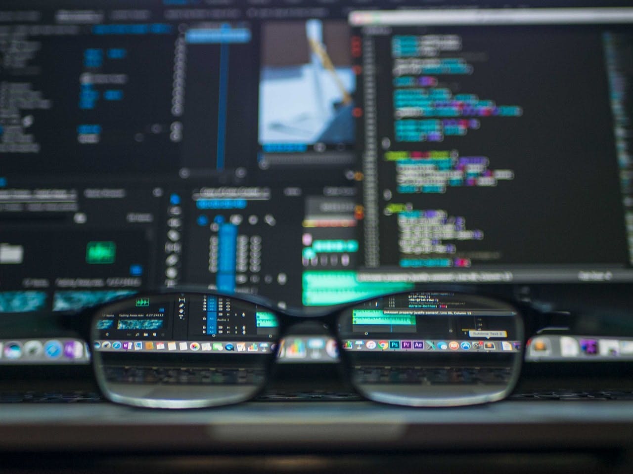 Glasses rest on a laptop keyboard, with computer screens displaying colorful programming code and software interfaces in the background, giving a blurred effect through the lenses.