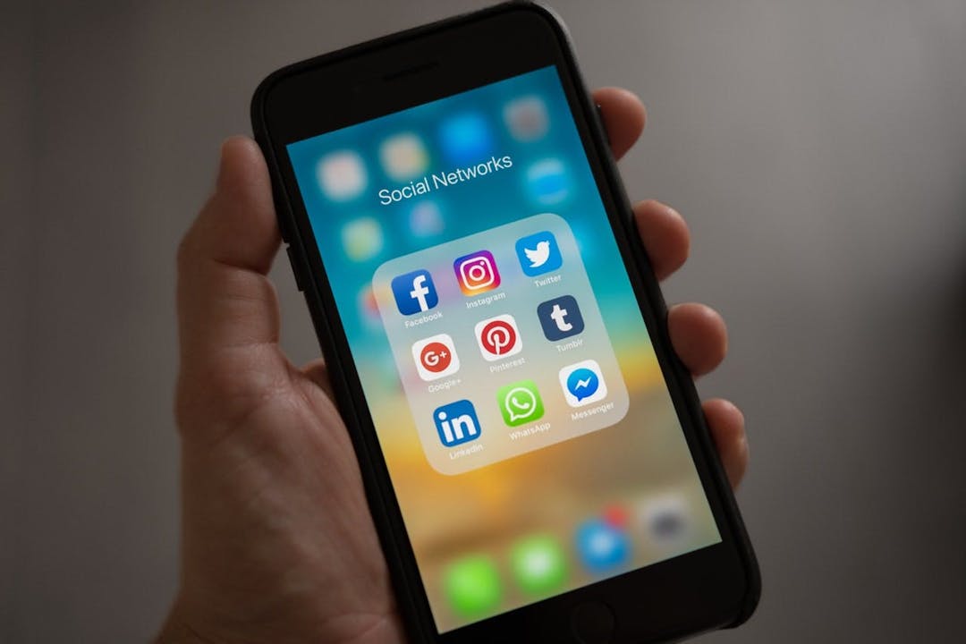 A hand holds a smartphone displaying a folder titled "Social Networks" with app icons: Facebook, Instagram, Twitter, Google+, Pinterest, Tumblr, LinkedIn, WhatsApp, and Messenger, against a blurred background.