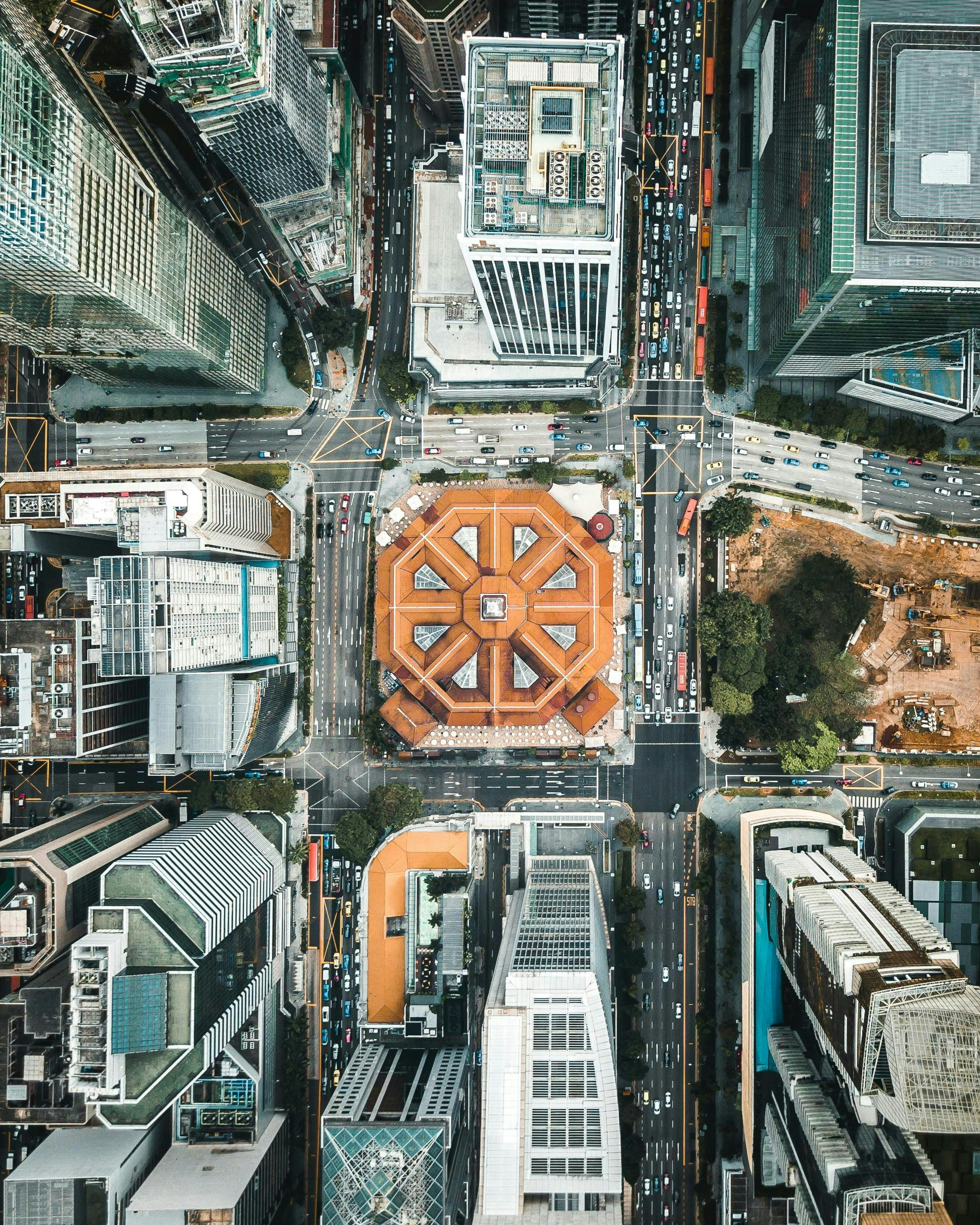 Aerial view of a city intersection with a central octagonal building. Surrounding skyscrapers tower above busy streets lined with cars and a construction site visible at the edge.