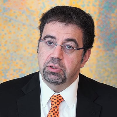 A man in a black suit and orange tie speaks against a colorful, abstract background. He has glasses and short, dark hair. 

[No text found]