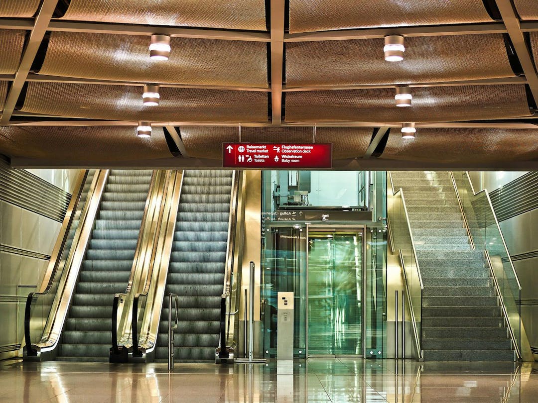 An escalator ascends on the left, an elevator has closed glass doors in the center, and a staircase rises on the right, in a brightly-lit, modern indoor setting. 

Text: "Restaurants, Toilets, Services".