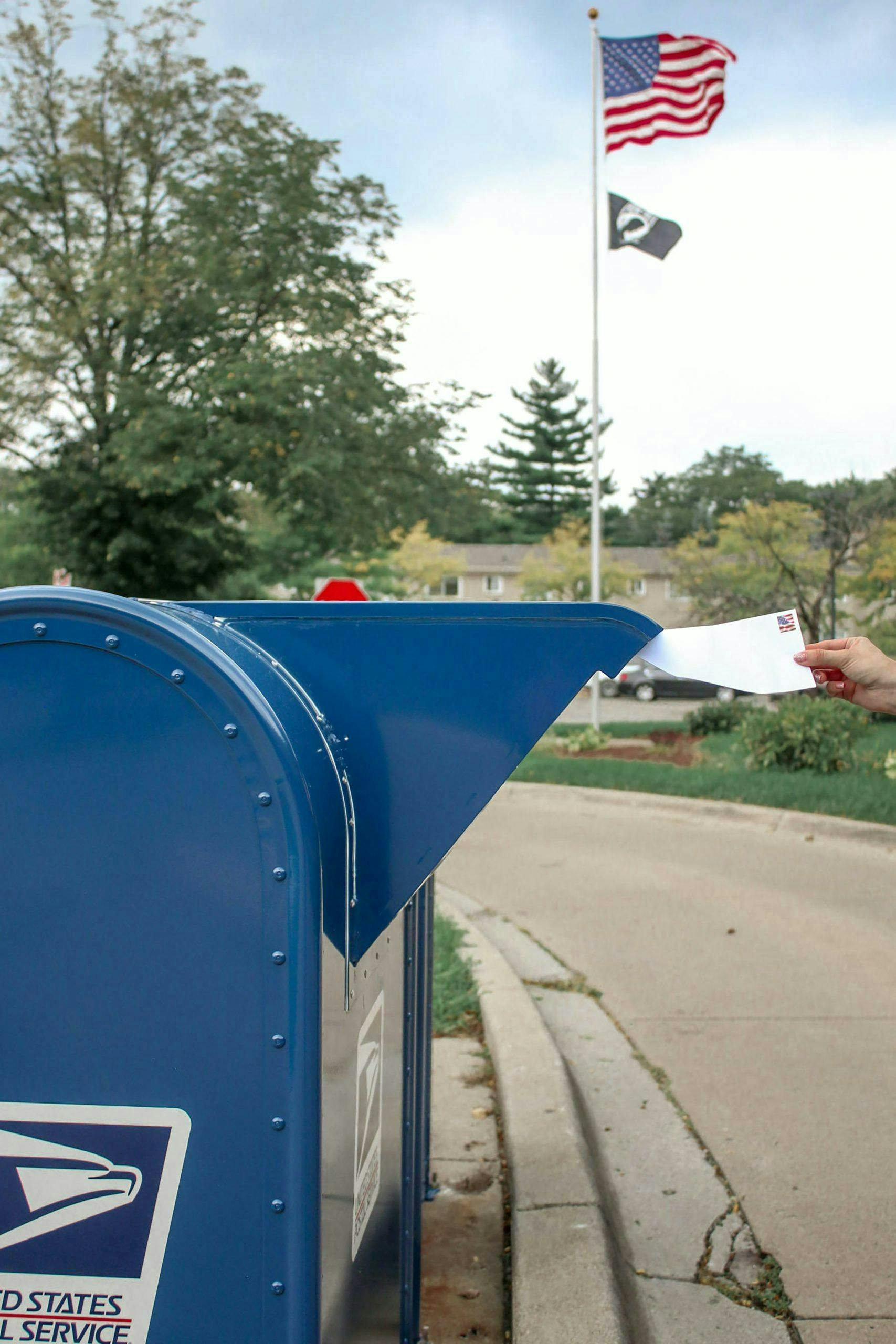 A person is placing a letter into a blue United States Postal Service mailbox on a suburban street, with trees and a U.S. flag visible in the background.
