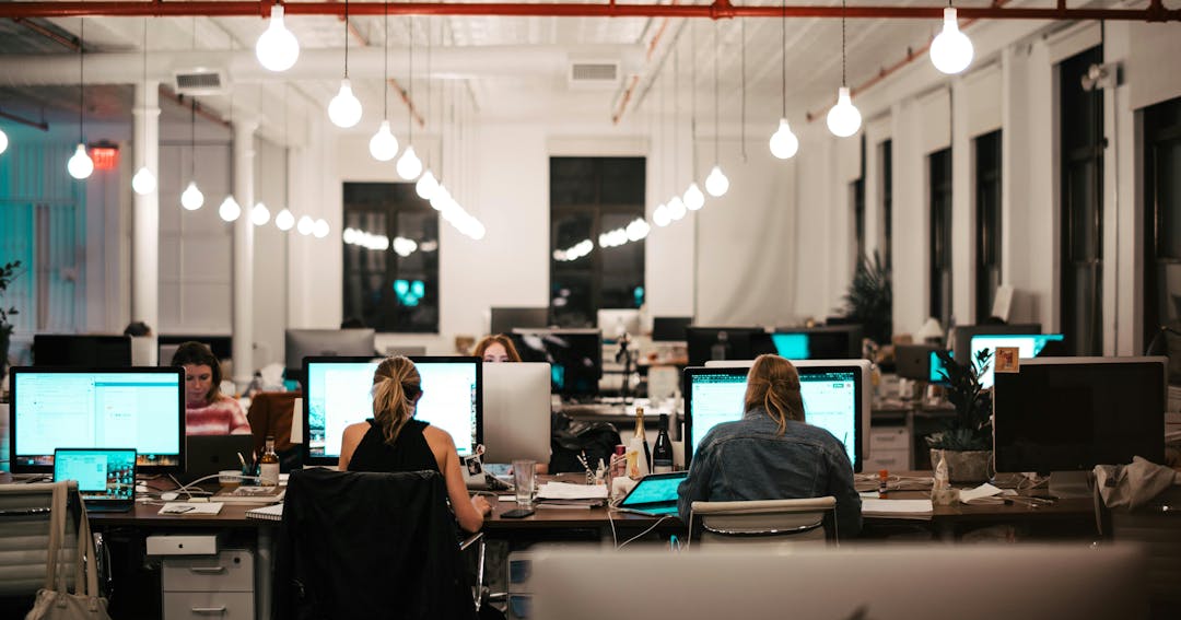 People work at desks surrounded by computers in a brightly lit, open-plan office with white walls and large windows. Hanging lights create a modern atmosphere.