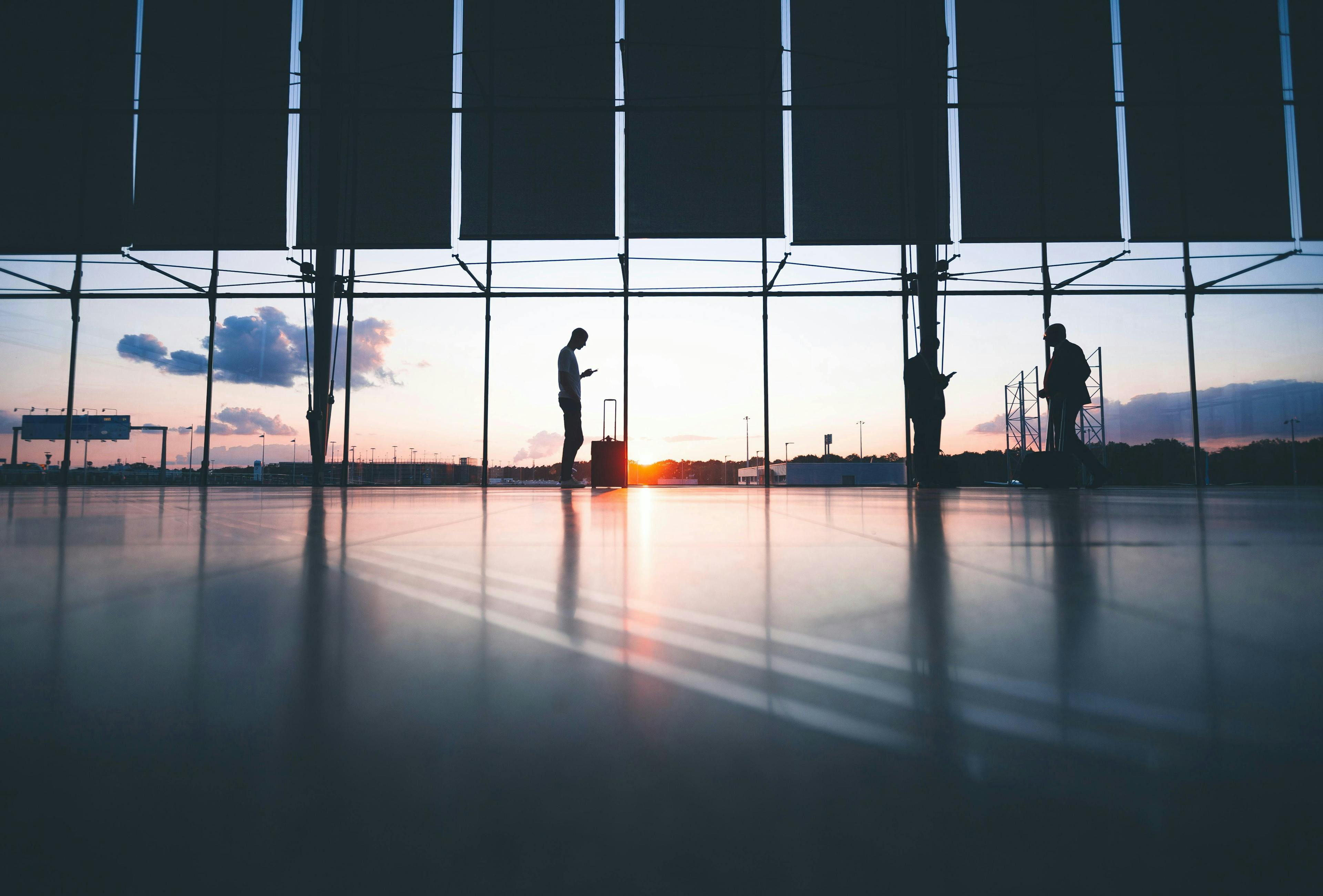 Silhouetted individuals stand and interact with their devices near large windows at an airport terminal during a scenic sunset with scattered clouds.