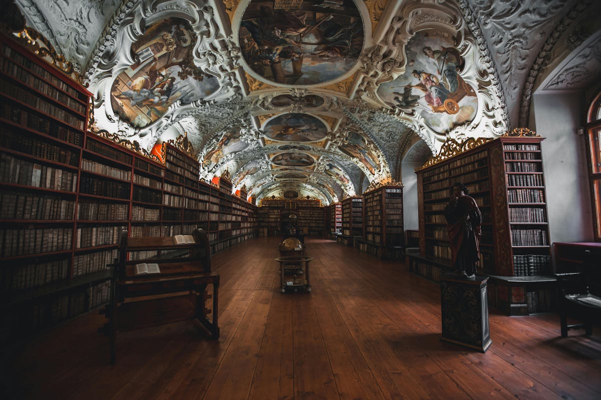 Library with ornate, painted ceilings; wooden floors and shelves lined with old books; a large globe in the center; statues and reading benches along the walls.