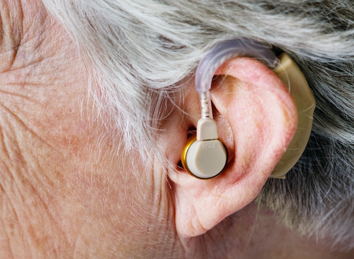 A hearing aid sits in the ear of an older person with grey hair and wrinkled skin, highlighting the close-up detail of the ear and hair.